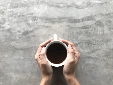 woman holding a white cup of hot black Americano coffee with her both hands on viva board or cement board table in coffee shop with copy space on top of frameの写真素材