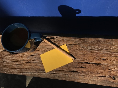 hot black coffeeâs put on natural wood bench with blank yellow note paper and brown pencil, and its shadow reflects on navy blue wall in present morningの写真素材