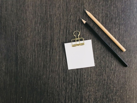 white note paper with golden clip on it and black and brown pencils put on dark brown table with copy space on left side of frameの写真素材