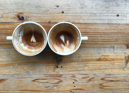 two white cups of finished latte coffee with flower and heart shape latte art left in cups on light brown wooden tableの写真素材