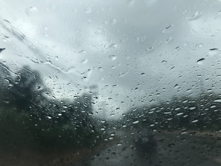 rain drops on car's windscreen with blurred stormy gray sky, hardly to see a car in front, and trees along the roadの写真素材