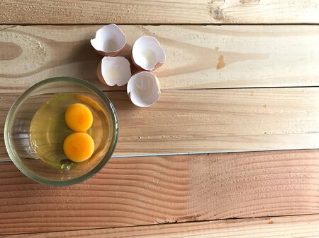 studio top view shot of two fresh yolk and egg white in clear glass cup and four eggshells beside it on natural rubber wood board with copy space on right side of frame