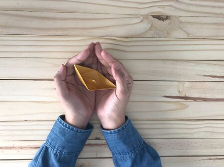 man wears blue jeans shirt is holding a small yellow paper boat origami in hands with light brown rubber softwood table on backgroundの写真素材