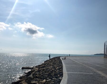 two men are fishing at the rocky shore with sunray on very bright blue sky, twinkle blue sea and...