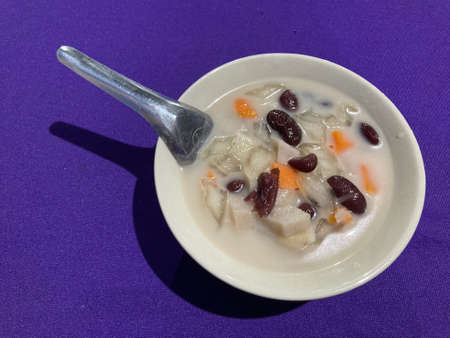 bowl of Tubo which is a dessert of Southern Thailand that has red beans, taro, sweet potato, pumpkin, jelly, coconut milk, sugar and salt as an ingredient, put on purple table with a silver spoonの写真素材