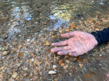 an Asian woman's hand with a black long sleeve swimming suit put in a bright crystal clear river with small brown pebbles beneath itの写真素材