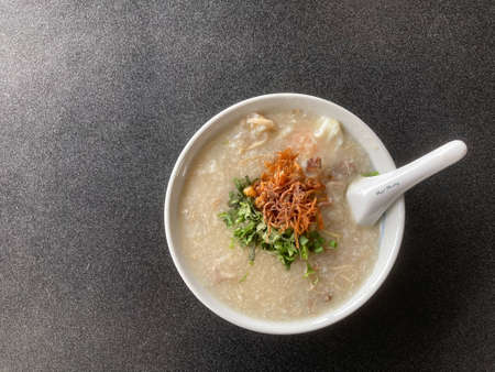 a bowl of rice congee with minced pork, boiled egg topped with fried shallot and fresh coriander, and white porcelain spoon put on a gray tableの写真素材
