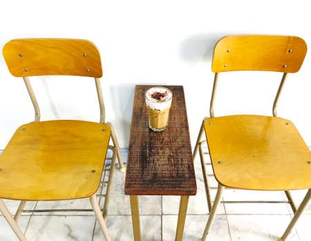 a glass of iced cappuccino coffee on a natural brown rectangle wooden table and two school chairs with a white wall on the backgroundの写真素材