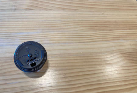 a takeaway black cup of hot coffee is put at the left of a wave-pattern light brown wooden tableの写真素材