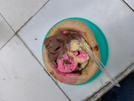 Three scoops of chocolate, pink, and yellow ice cream melt inside a halved coconut shell, served with a plastic spoon on a bright white tiled surface. A delicious and refreshing tropical dessert.の写真素材