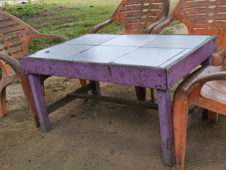 A weathered purple wooden table with a tiled top is surrounded by worn orange plastic chairs on sandy ground. Green grass is visible in the background, creating a rustic outdoor scene.の写真素材