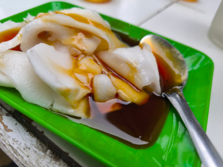 A close-up view of a sweet, translucent white dessert or snack drenched in rich brown syrup, served on a bright green plate with a spoon.の写真素材