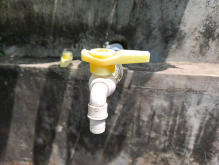 A white plastic water tap with a yellow lever handle is mounted on a rough, dark concrete wall, illuminated by natural sunlight.の写真素材