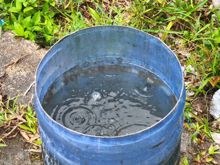 A blue metal barrel, partially submerged in the ground, collects water with visible ripples and a prominent bubble on its surface, surrounded by green foliage.の写真素材