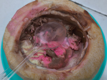 Delicious pink and chocolate ice cream melting inside a fresh, halved coconut, ready to be enjoyed with a plastic spoon.の写真素材