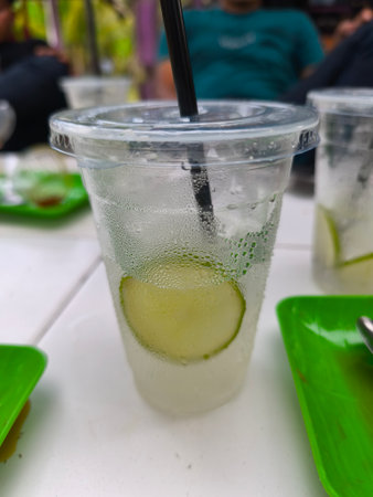 A close-up view of a refreshing lime-infused cold drink in a clear plastic cup with a black straw, showing condensation. It sits on a white table with blurred green plates and people in the background.の写真素材