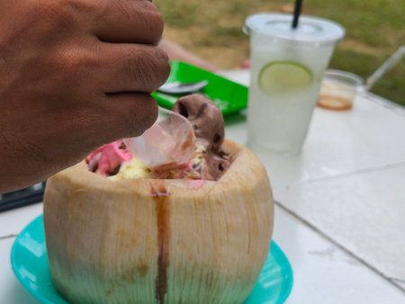 A persons hand scoops colorful, melting ice cream from a coconut shell, served outdoors on a sunny day with a refreshing drink.の写真素材