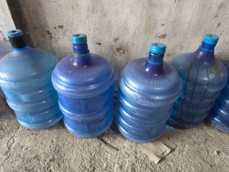 A lineup of empty, used blue plastic water gallons stands against a textured wall and rough concrete floor, reflecting a sense of utility and storage.の写真素材