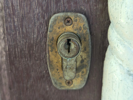 A close-up view of a worn, oxidized brass door lock cylinder embedded in a dark wooden door, showcasing its rustic texture.の写真素材