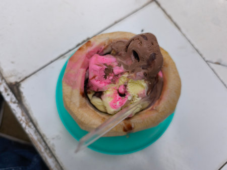 A refreshing tropical dessert featuring scoops of pink, vanilla, and chocolate ice cream served inside a natural coconut shell on a tiled surface.の写真素材