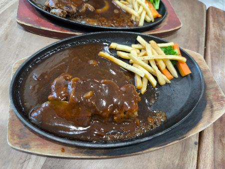 Close up of a sizzling hot plate featuring a hearty meal of steak, rich gravy, crispy french fries, and steamed vegetables on a wooden boardの写真素材