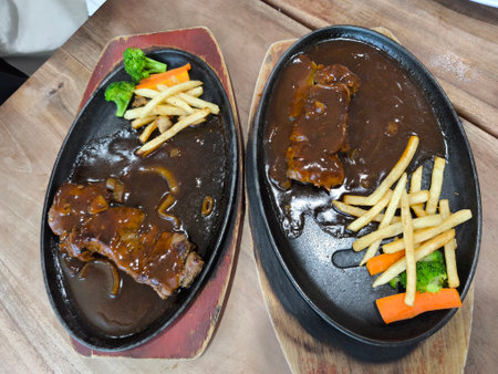 Two sizzling steaks are served on hot cast iron platters with rich brown gravy, accompanied by crispy french fries, fresh broccoli, and sweet carrots, presented on a wooden tableの写真素材