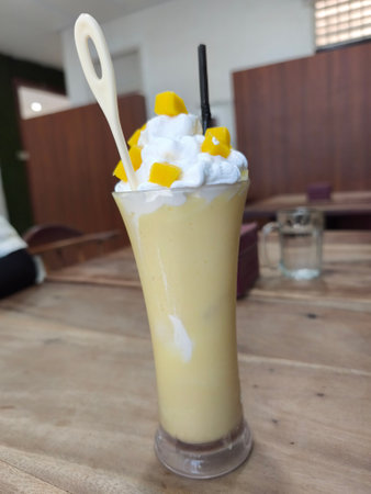 Delicious mango smoothie in a tall glass, garnished with fluffy whipped cream and vibrant mango chunks, served with a spoon and straw on a rustic wooden tableの写真素材
