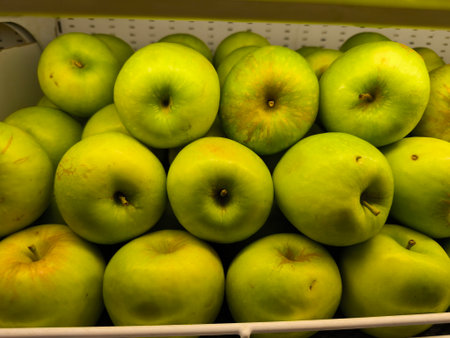 Fresh, bright green apples are arranged closely together on a white display shelf, showcasing their natural colors and textures, ready for purchaseの写真素材