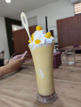 Enjoy a delightful mango smoothie, topped with luscious whipped cream and vibrant mango cubes, served in a tall glass on a rustic wooden tableの写真素材