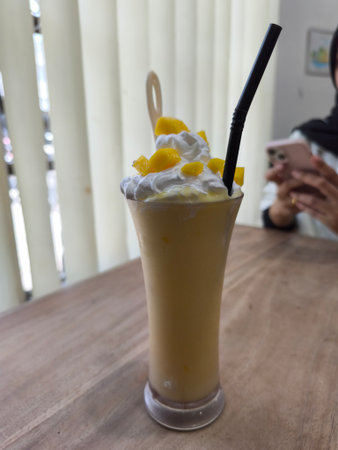 Refreshing mango smoothie in a tall glass, garnished with whipped cream and fresh mango cubes on a wooden tableの写真素材