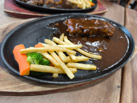 Delicious sizzling steak served on a hot plate, accompanied by crispy french fries, steamed carrots, and broccoli, presented on a wooden boardの写真素材