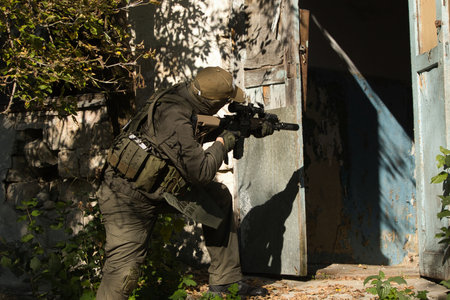 Tactical operator in full combat gear including plate carrier, tactical pants, and cap, holding rifle with optics in urban training environmentの写真素材
