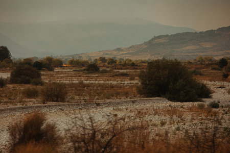 A dry rocky plain with scattered bushes and hazy mountain ridges in the distance, Greece, 16 August 2025.の写真素材