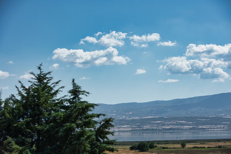 Wide panoramic view of a calm lake and rolling hills under a blue sky with scattered clouds,...