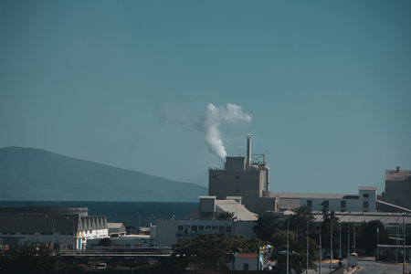 Industrial factory complex with smokestacks emitting steam and smoke against a clear blue sky, viewed from the roadside during a summer journey. Heavy industry August 29, 2025.のeditorial素材