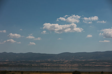 Wide panoramic view of a calm lake and rolling hills under a blue sky with scattered clouds, photographed during a summer road trip in August 2025の写真素材