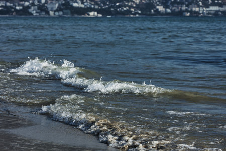 Vlora, Albania â August 2025: Dynamic coastal waves roll towards the shore with the seaside city and mountain landscape of Vlora.の写真素材