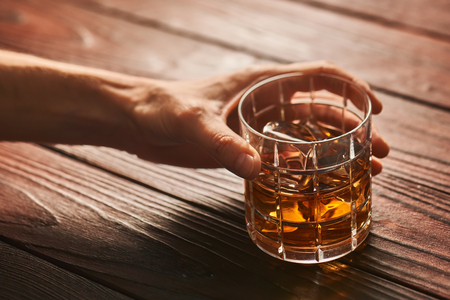 Man's hand holding Glass of whiskey with ice cubes on rustic wooden table with copy-spaceの写真素材