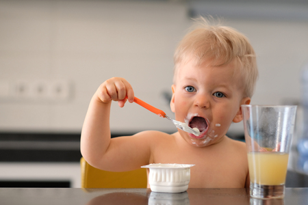 Adorable one year old baby boy eating yoghurt with spoon. Dirty messy face of toddler child.の写真素材