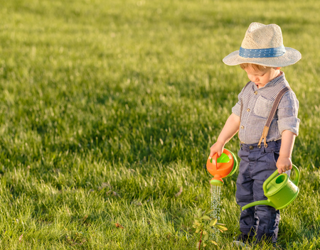 Portrait of toddler child outdoors. Rural scene with one year old baby boy wearing straw hat using watering canの写真素材