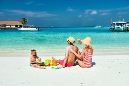 Family on beach, young couple with three year old boy. Woman applying sun screen protection lotion on man's back. Summer vacation at Maldives.の写真素材