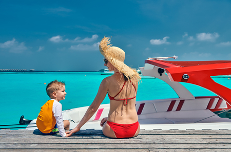 Three year old toddler boy with mother sitting on wooden jetty with boat. Summer family vacation at Maldives.の写真素材