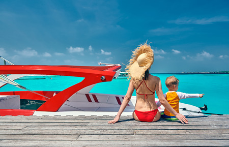 Three year old toddler boy with mother sitting on wooden jetty with boat. Summer family vacation at Maldives.の写真素材