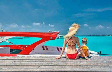 Three year old toddler boy with mother sitting on wooden jetty with boat. Summer family vacation at Maldives.の写真素材