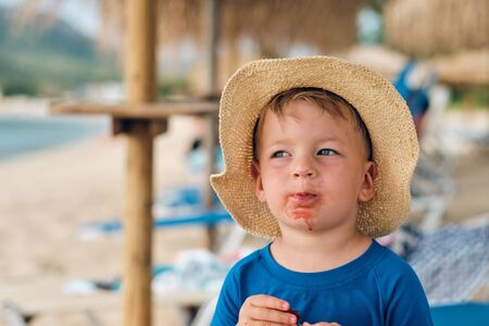 Two year old toddler boy on beach eating fruit. Dirty messy face of happy kid.の写真素材