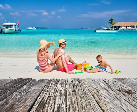 Family on beach, young couple with three year old boy. Woman applying sun screen protection lotion on man's back. Summer vacation at Maldives.の写真素材