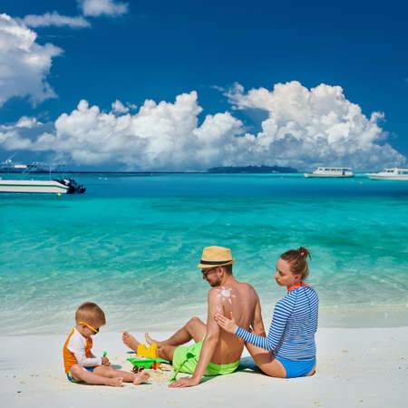Family on beach, young couple with three year old boy. Woman applying sun screen protection lotion on man's back. Summer vacation at Maldives.の写真素材
