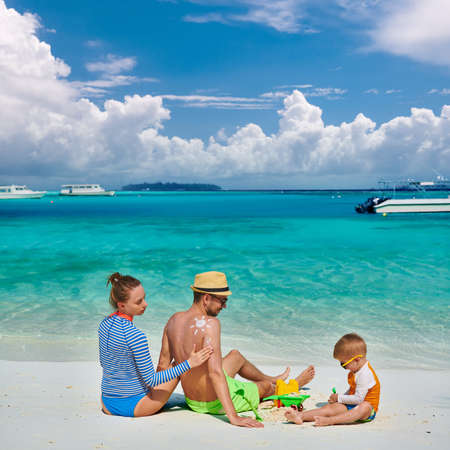 Family on beach, young couple with three year old boy. Woman applying sun screen protection lotion on man's back. Summer vacation at Maldives.の写真素材
