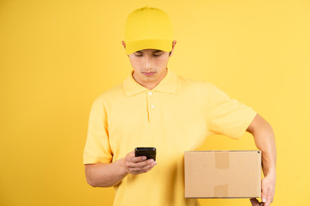 Portrait of delivery male holding cargo box and using phone on yellow backgroundの写真素材