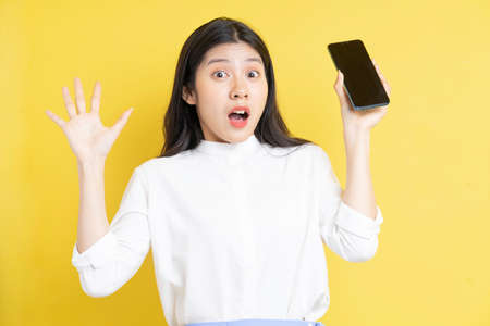 Young asian girl holding phone with expression on yellow backgroundの写真素材